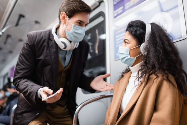 man in headphones holding smartphone and gesturing near african american woman in medical mask in subway - Фото, изображение