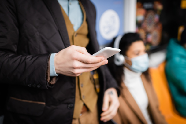 мужчина держит смартфон в метро рядом с африканской американкой в медицинской маске на размытом фоне - Фото, изображение