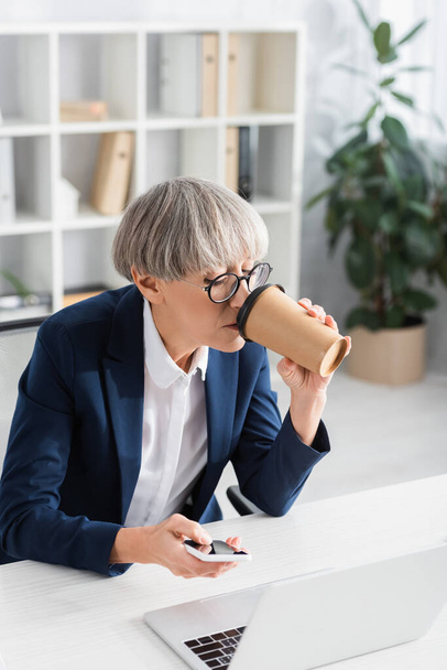 лидер команды средних лет в очках, пьющий кофе, чтобы пойти во время использования смартфона рядом с ноутбуком на столе - Фото, изображение