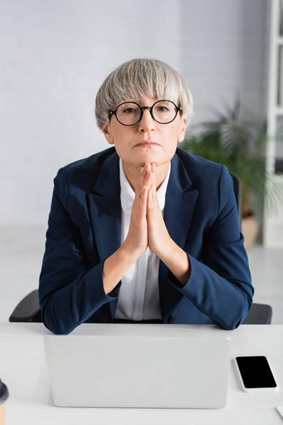 middle aged team leader in glasses sitting with praying hands near gadgets - Photo, Image