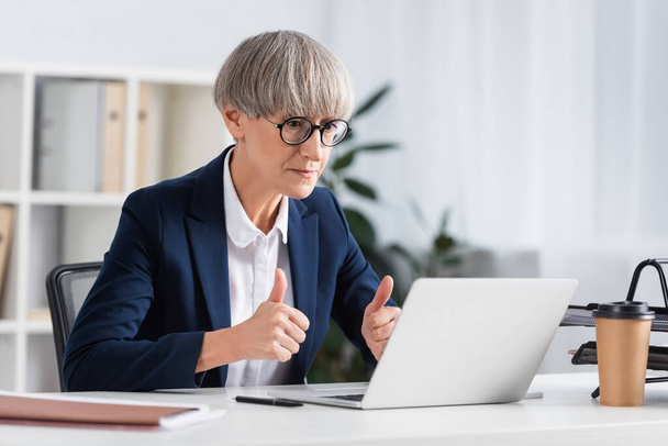 mature team leader in glasses showing thumbs up while looking at laptop  - Photo, Image