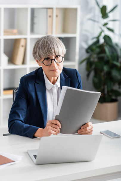 middle aged team leader in glasses holding folder near gadgets on table - Photo, Image