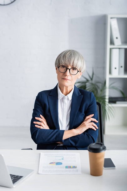 femme d'affaires d'âge moyen dans des lunettes assis avec les bras croisés près des cartes et des graphiques sur le bureau - Photo, image