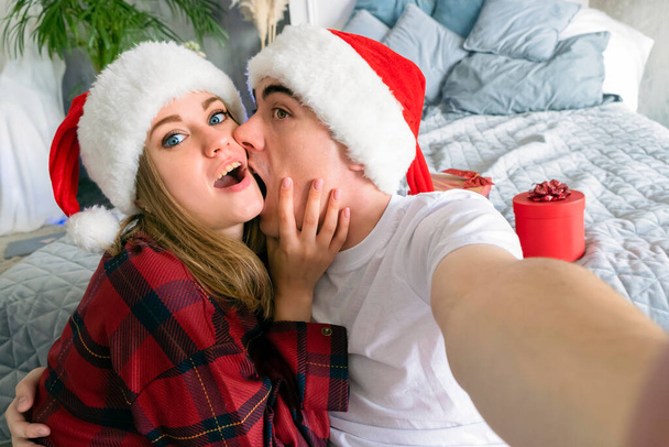 Fiesta de Navidad en línea. Hombre mordiendo mujer tomando selfie usando sombreros de santa y pijamas rojos en el fondo de los regalos de Año Nuevo - Foto, Imagen