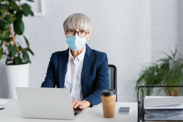 middle aged team leader in glasses and medical mask using laptop near smartphone and paper cup on desk - Фото, изображение