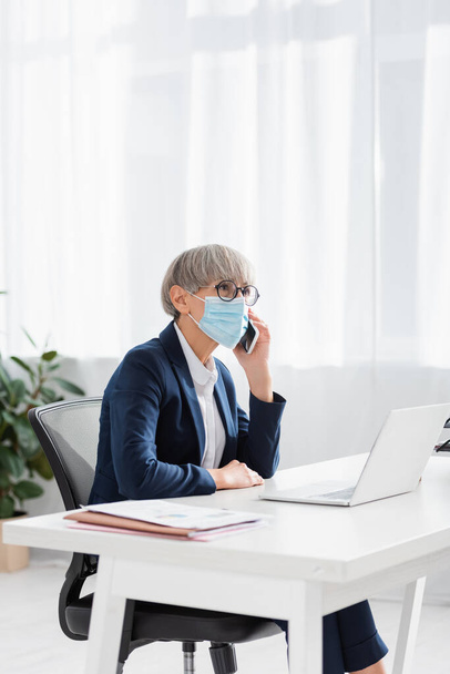 leader del team di mezza età in occhiali e maschera medica che parla sullo smartphone vicino al laptop sulla scrivania - Foto, immagini