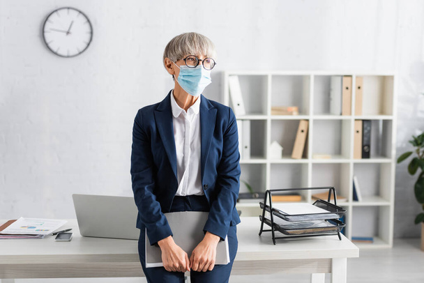 mature team leader in glasses and medical mask holding folder while standing near workplace - Фото, зображення