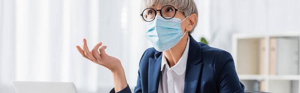 mature team leader in glasses and medical mask gesturing while looking up in office, banner - Foto, Imagem