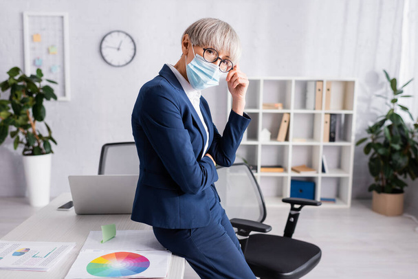 Reifer Teamleiter mit Brille und medizinischer Maske lehnt auf Stuhl neben Schreibtisch - Foto, Bild