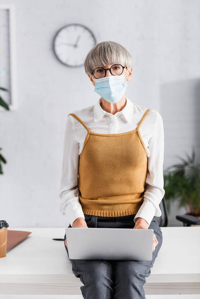 középkorú csoportvezető szemüvegben és orvosi maszkban laptoppal, miközben az irodában ül - Fotó, kép