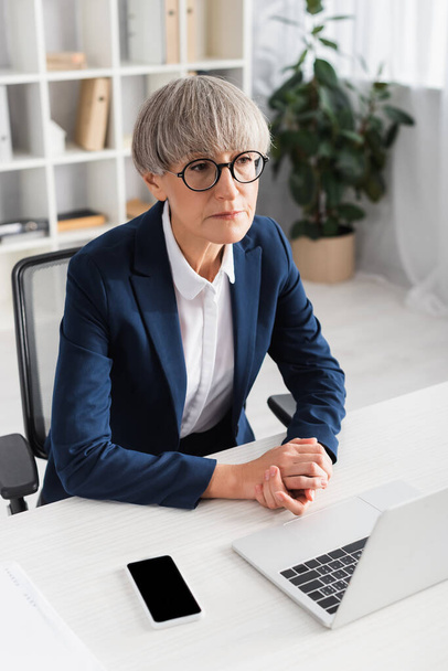middle aged team leader in glasses sitting with clenched hands near devices on desk - Фото, изображение