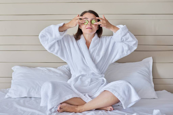 Mature female in bathrobe doing home spa treatments with cucumbers - Zdjęcie, obraz