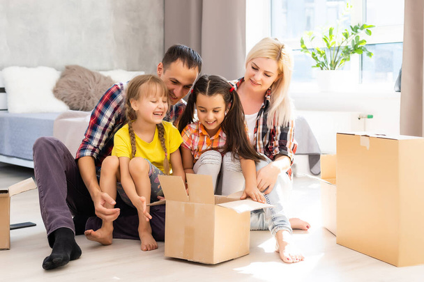 Kaukasische Familie, Mann, Frau und zwei Mädchen sitzen auf dem Fußboden, packen Kisten aus und lächeln im neuen Haus. Hinter ihnen Umzugskartons. - Foto, Bild