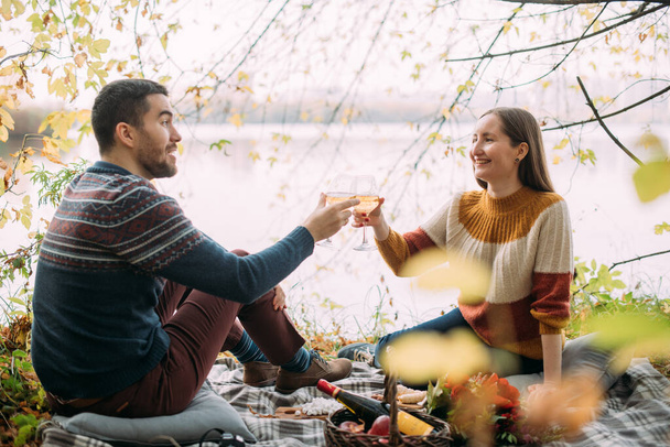 Een liefdevol koppel op een date tijdens een picknick op het meer. Jongeman en vrouw zitten in de herfst op een deken met wijn en fruit in de buurt van het water in het park. Een jong gezin viert een jubileum. - Foto, afbeelding