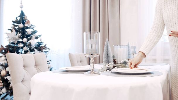 Een meisje in een witte jurk serveert borden aan de kersttafel. Feestelijke tafelschikking - Foto, afbeelding