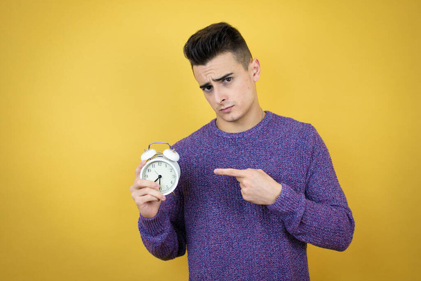 joven caucásico hombre sobre aislado amarillo fondo serio celebración y apuntando un reloj - Foto, imagen
