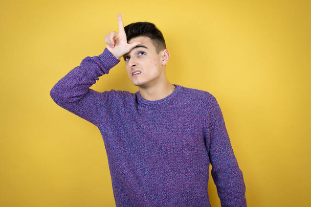 Young caucasian man over isolated yellow background making fun of people with fingers on forehead doing loser gesture mocking and insulting. - Photo, Image