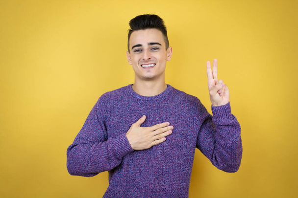 Jeune homme caucasien sur fond jaune isolé souriant jurer avec la main sur la poitrine et les doigts vers le haut, faisant un serment de fidélité promesse - Photo, image