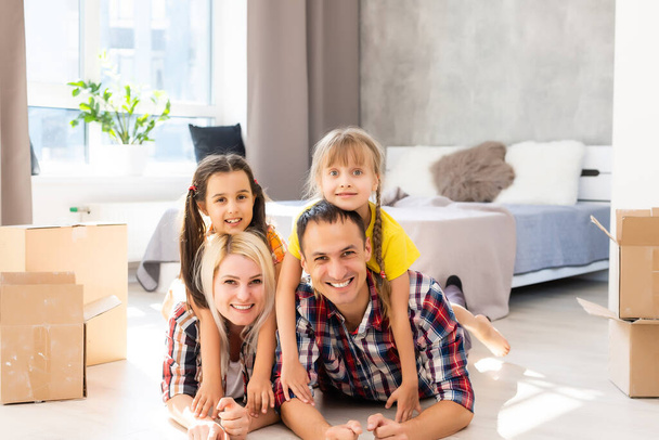 Caucasian family, man, woman and two girls sit on floor, unpack boxes and smile in new house. Behind them moving boxes. - Foto, afbeelding