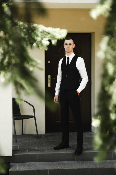 a guy dressed in suit pants and a shirt on top of which a classic vest stands near his house. the Butler. house attendants. stylish clothes of the young groom - Fotó, kép