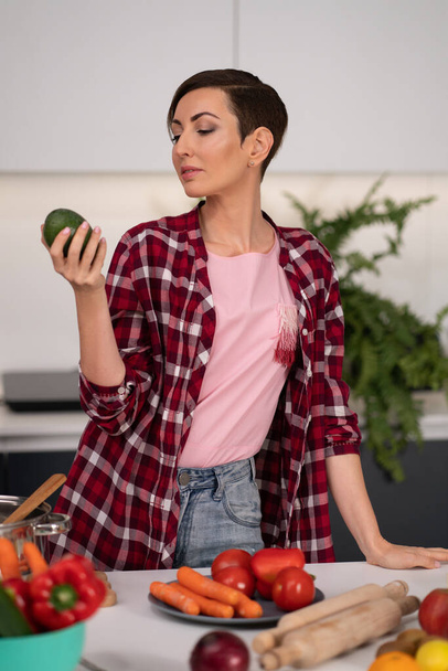 Looking at avocado and thinking pretty housewife with a short hairstyle prepares food in the kitchen. Healthy food at home. Healthy food leaving - vegan concept - Foto, immagini