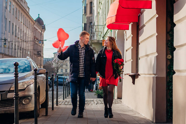 Valentines day couple walking outdoors. Man and woman hold hands with bouquet of red roses flowers and balloons on street. Romantic date - Zdjęcie, obraz
