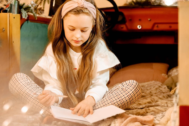 bonito bela menina de cabelos compridos escrevendo uma carta para Papai Noel. Atmosfera de Ano Novo, pede um desejo. a criança senta-se no carro e lê um boo - Foto, Imagem