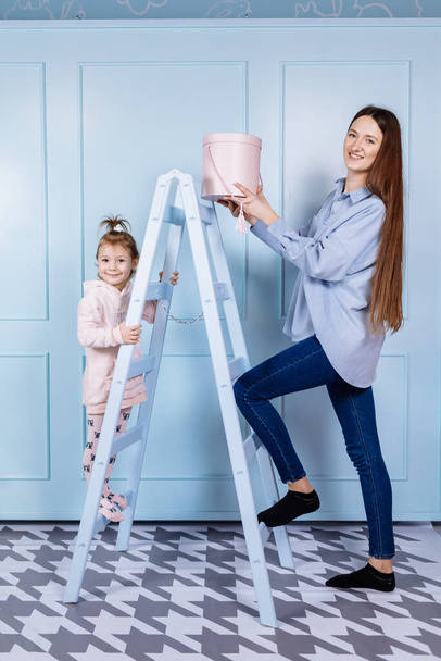 Glücklich liebende Familie. junge Mutter in blau und ihre kleine Tochter in pinkfarbener Kleidung spielen mit rosa Geschenkbox und blauer Leiter auf blauem Wandhintergrund. Glückliche Mütter, Babytag - Foto, Bild