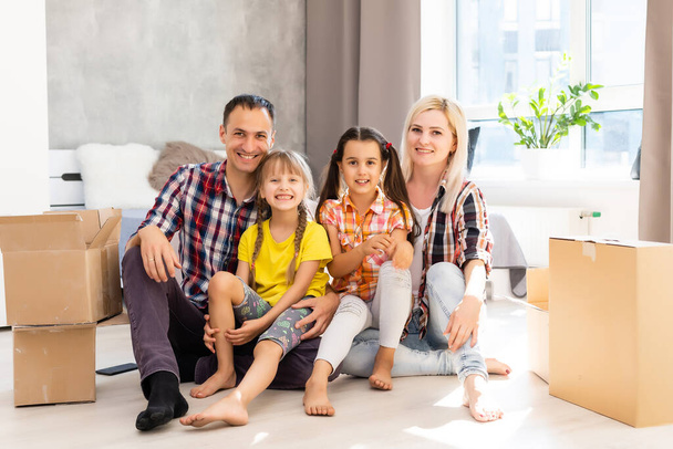 Família caucasiana, homem, mulher e duas meninas sentam-se no chão, desempacotam caixas e sorriem na nova casa. Atrás deles caixas móveis. - Foto, Imagem