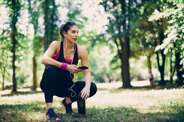 Jonge aantrekkelijke fit Kaukasische sportvrouw hurken in de natuur en met behulp van smart phone om muziek te zoeken voor hardlopen. - Foto, afbeelding