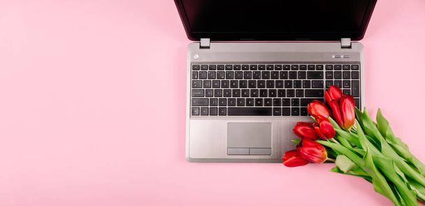 職場のノートパソコンとピンクの紙の背景に赤いチューリップの花束。テキストのコピースペースを持つブログの広告や可視化のためのテンプレート。名刺だ。休日証明書。バナー. - 写真・画像