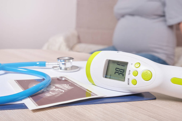 ultrasound picture, stethoscope and non-contact infrared thermometer with high temperature on the table, pregnant woman on the background - Fotoğraf, Görsel