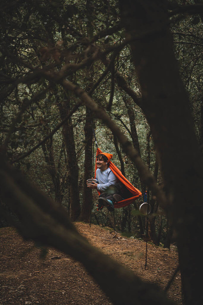 Hammock on trees in the forest, man lying in a hammock in the middle of a dark forest, orange hammock, man wears teal sweater and orange hat - Photo, Image