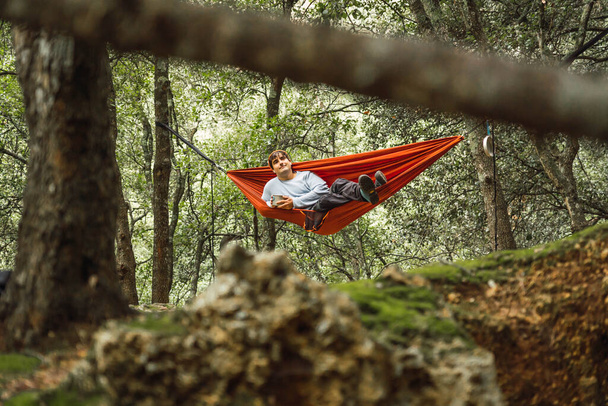 Hangmat aan bomen in het bos, man liggend in een hangmat in het midden van een donker bos, oranje hangmat, man draagt teal trui en oranje hoed - Foto, afbeelding