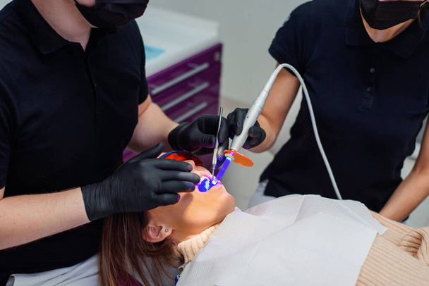 Female patient in bib and orange UV protective glasses in a dental clinic. Dentist in gloves using UV lamp. - Foto, afbeelding