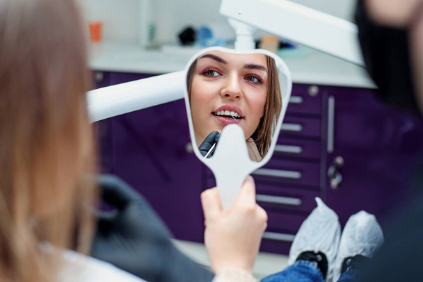Young woman patient looking at mirror and admires her new smile after dental treatment in the dentistry clinic. - Фото, зображення