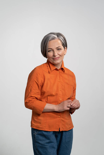 Happy middle aged woman looking at camera in orange shirt and denim pants. Pretty mid aged grey haired woman in orange shirt isolated on grey background - Fotografie, Obrázek