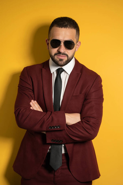 Serious handsome man in burgundy color suit and sunglasses looking at camera with hands folded isolated on yellow background - Foto, imagen