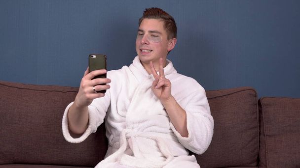 Un joven con manchas en la cara se toma una selfie. Parches de hidrogel azul para el rejuvenecimiento facial. Un hombre sentado en un abrigo blanco en el sofá. - Foto, imagen