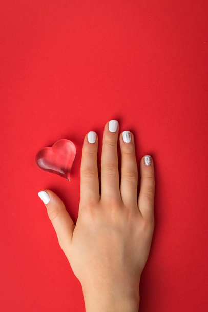 Ένα γυναικείο χέρι με λευκό μακιγιάζ και γυάλινη καρδιά σε κόκκινο φόντο. Ένα σύμβολο αγάπης και ζωής. - Φωτογραφία, εικόνα