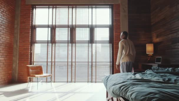 African American man getting out of bed, walking to floor to ceiling window with blinds, stretching and using smartphone after waking up in the morning at home - Filmati, video