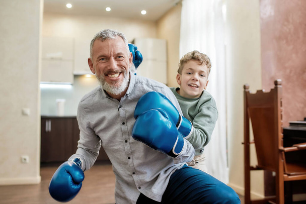 Portrait of a cheerful grandpa and little boy grandson wearing blue boxing gloves having fun and smiling at camera while having boxing training - Valokuva, kuva