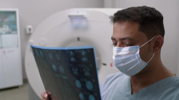 PAN shot van Arabische mannelijke arts in het gezicht masker staan in radiologie kamer en het bestuderen van MRI scan van de patiënt, dan kijken naar camera - Video