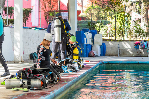 žena připravuje potápěčské vybavení na okraji bazénu, zatímco ostatní muži v pozadí jsou již na sobě zařízení - Fotografie, Obrázek