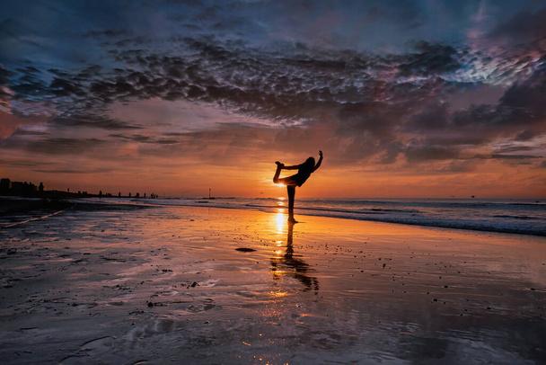 Σιλουέτα μιας νεαρής γυναίκας που κάνει γιόγκα στην παραλία κατά τη διάρκεια ενός όμορφου χρυσαφένιου, μωβ ηλιοβασιλέματος - Φωτογραφία, εικόνα