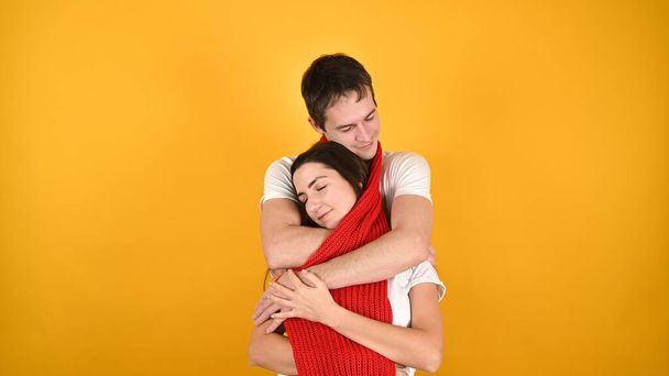 Les jeunes couples aimants se sont enveloppés dans une longue écharpe rouge chaud. Jeune homme et femme câlins sur fond jaune avec espace de copie. Concept de Saint-Valentin.  - Photo, image