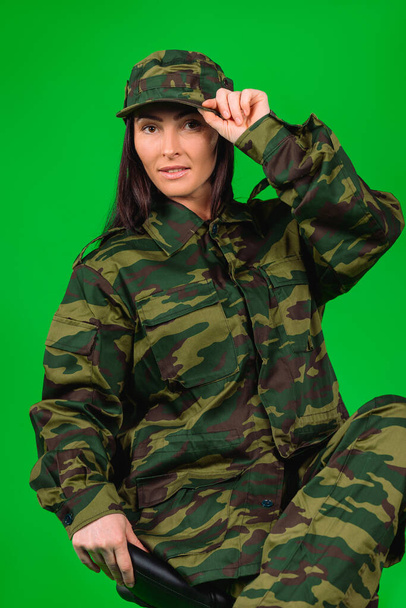 Young attractive woman in khaki military uniform sits on black chair posing on a green background. High quality photo - Fotografie, Obrázek