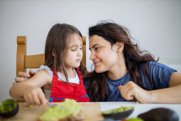 Gelukkige moeder en dochter aan een tafel maken avocado toast - Foto, afbeelding