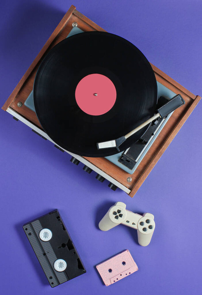 Flat lay éléments rétro sur fond violet. Lecteur vinyle, vidéo, cassette audio, manette de jeu. Culture pop, années 80. Vue du dessus - Photo, image