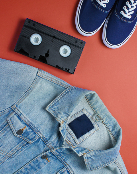 Retro style stuff. Videotape, jeans jacket, sneakers on red background. Top view - Photo, image